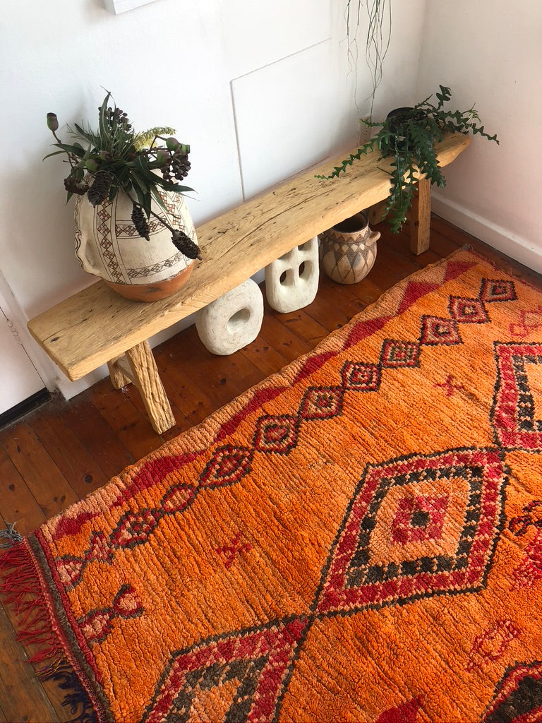 An orange and red rug on the floor with a diamond pattern, next to a wooden bench with two pots resting on it.