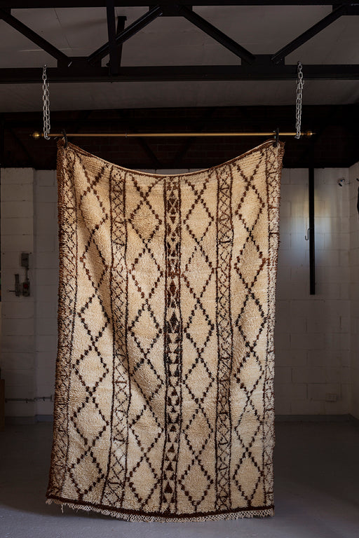 A very large cream rug with a brown diamond pattern, hanging from the ceiling by a pole and two chains.